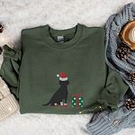 Military Green crewneck sweatshirt laid flat with a knit blanket and pine cones, embroidered with a sitting black Labrador in a Santa hat and red booties beside a green gift box