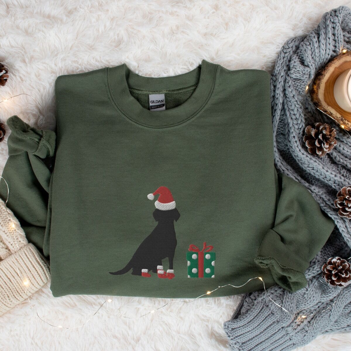 Black Lab Christmas Sweatshirt – Military Green Military Green crewneck sweatshirt laid flat with a knit blanket and pine cones, embroidered with a sitting black Labrador in a Santa hat and red booties beside a green gift box