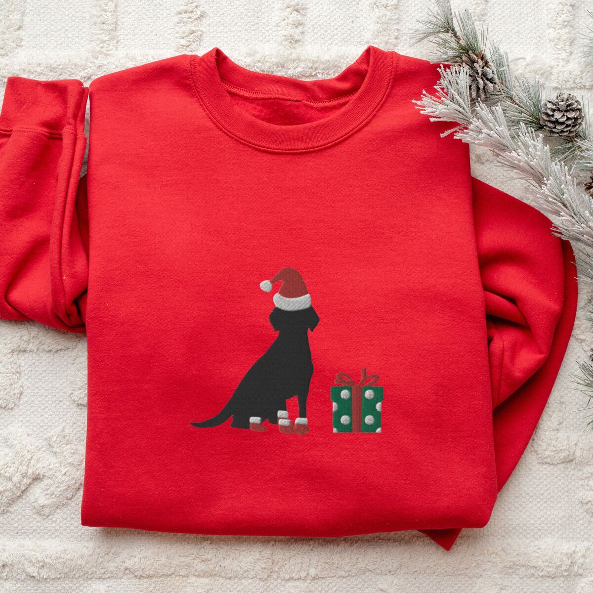Black Lab Christmas Sweatshirt - Red Red crewneck sweatshirt folded on a white rug with frosted pine, embroidered with a sitting black Labrador in a Santa hat and red booties beside a green gift box