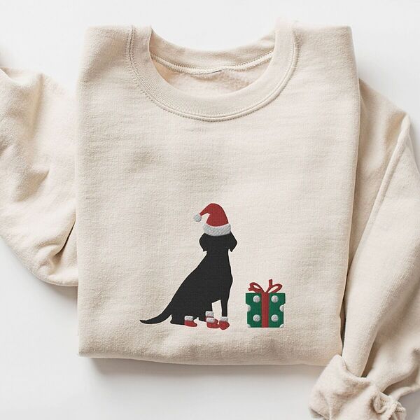 Sand crewneck sweatshirt laid flat with rolled cuffs, embroidered with a sitting black Labrador in a Santa hat and red booties beside a green gift box