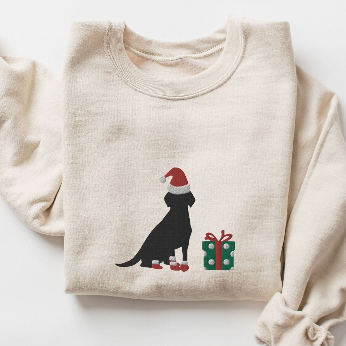 Black Lab Christmas Sweatshirt – Sand Sand crewneck sweatshirt laid flat with rolled cuffs, embroidered with a sitting black Labrador in a Santa hat and red booties beside a green gift box