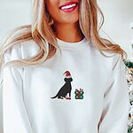 Woman smiling by a Christmas tree in a White crewneck sweatshirt embroidered with a sitting black Labrador in a Santa hat and red booties beside a green gift box