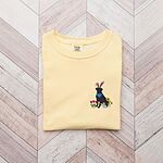 Butter T-shirt folded on a wood floor, embroidered with a sitting black lab in bunny ears and a blue bandana with pink tulips, yellow flowers and a little bunny on the left chest