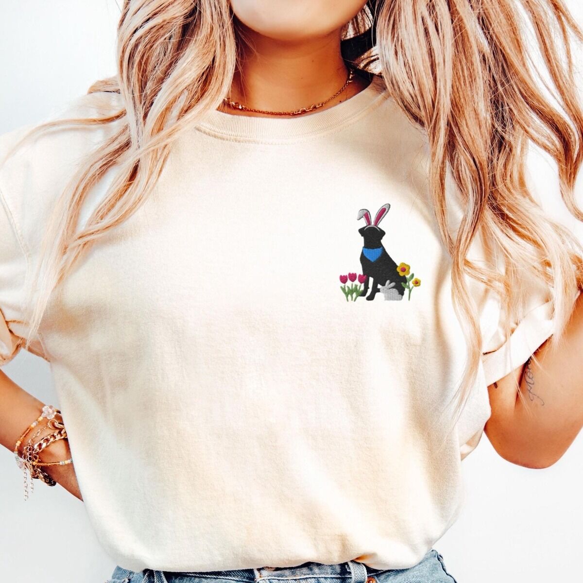 Black Lab Easter Shirt – Ivory Woman wearing a Ivory T-shirt embroidered with a sitting black lab in bunny ears and a blue bandana with pink tulips, yellow flowers and a little bunny on the left chest