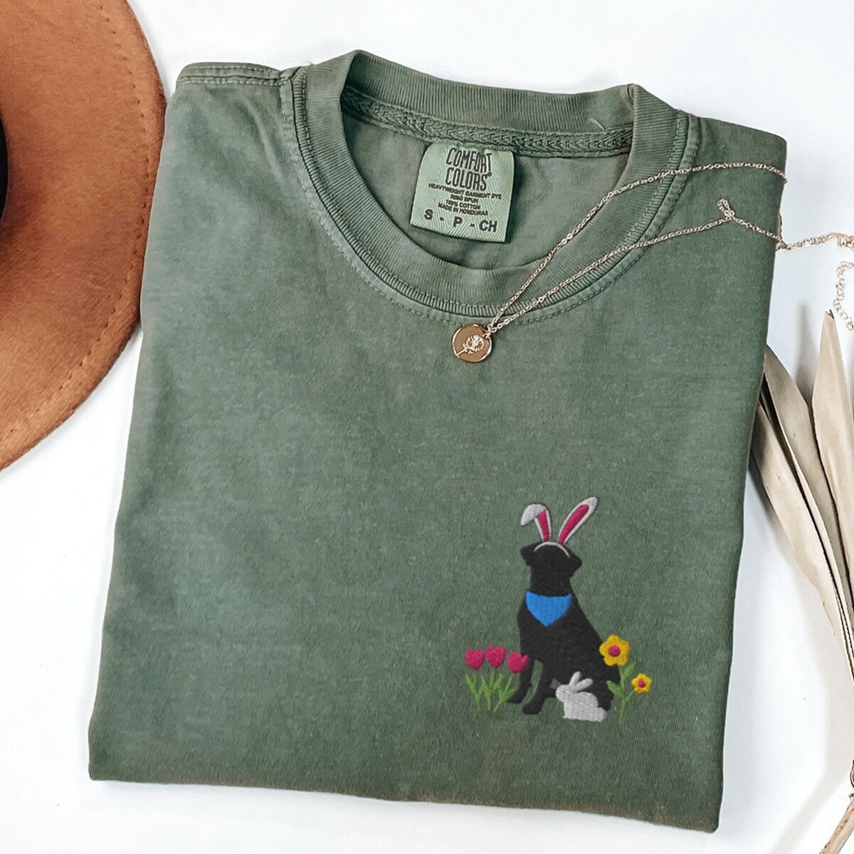 Black Lab Easter Shirt – Moss Moss T-shirt folded with a gold necklace, embroidered with a sitting black lab in bunny ears and a blue bandana with pink tulips, yellow flowers and a little bunny on the left chest