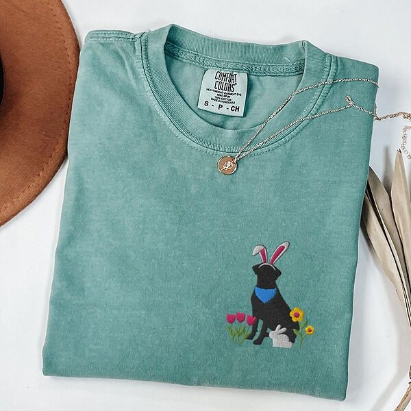 Seafoam T-shirt folded with a gold necklace, embroidered with a sitting black lab in bunny ears and a blue bandana with pink tulips, yellow flowers and a little bunny on the left chest