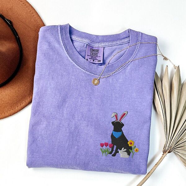 Violet T-shirt folded with a gold necklace, embroidered with a sitting black lab in bunny ears and a blue bandana with pink tulips, yellow flowers and a little bunny on the left chest