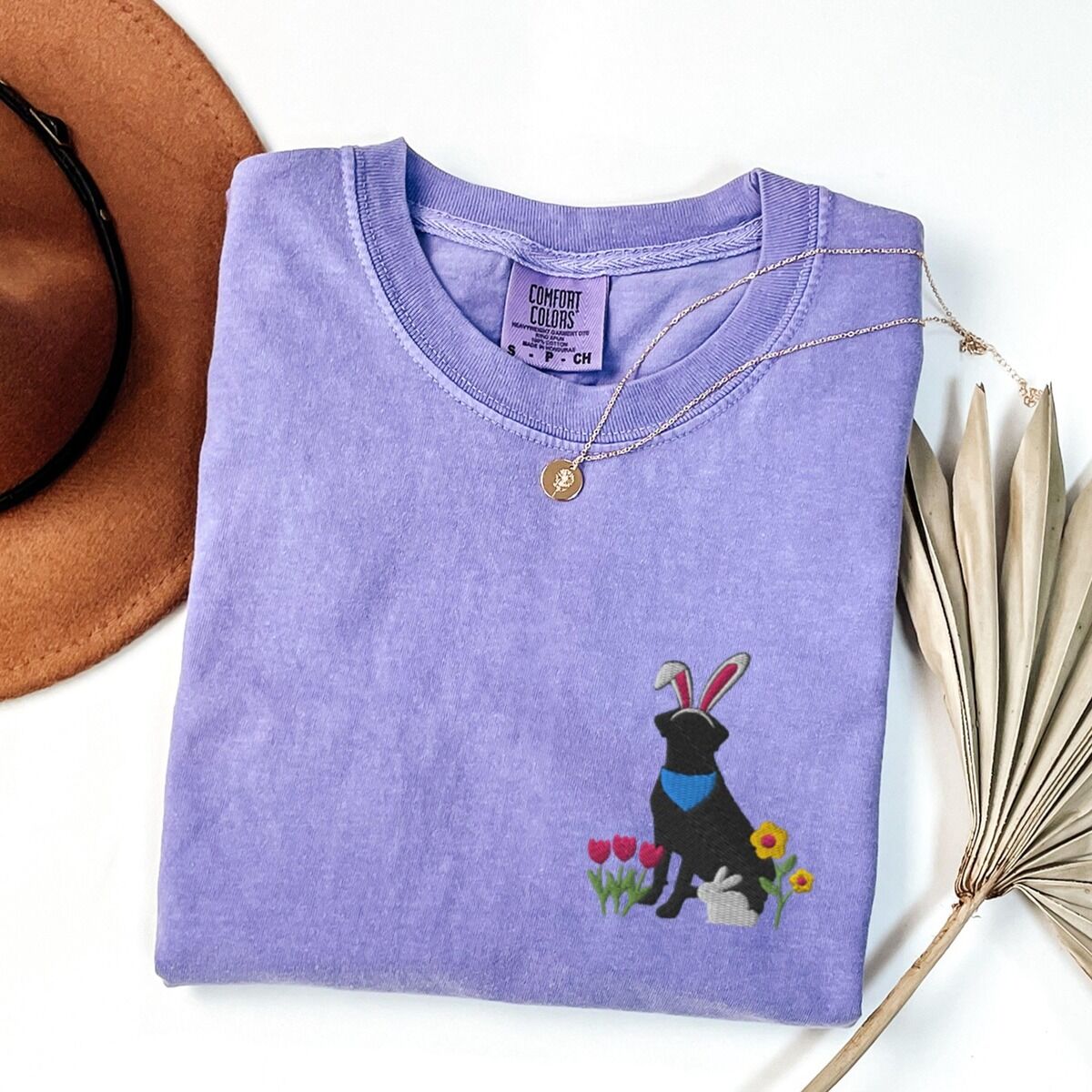 Black Lab Easter Shirt – Violet Violet T-shirt folded with a gold necklace, embroidered with a sitting black lab in bunny ears and a blue bandana with pink tulips, yellow flowers and a little bunny on the left chest