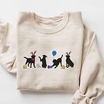 Sand crewneck sweatshirt laid flat, embroidered with four black lab silhouettes in bunny ears with a little bunny, Easter eggs, daffodils and a blue balloon