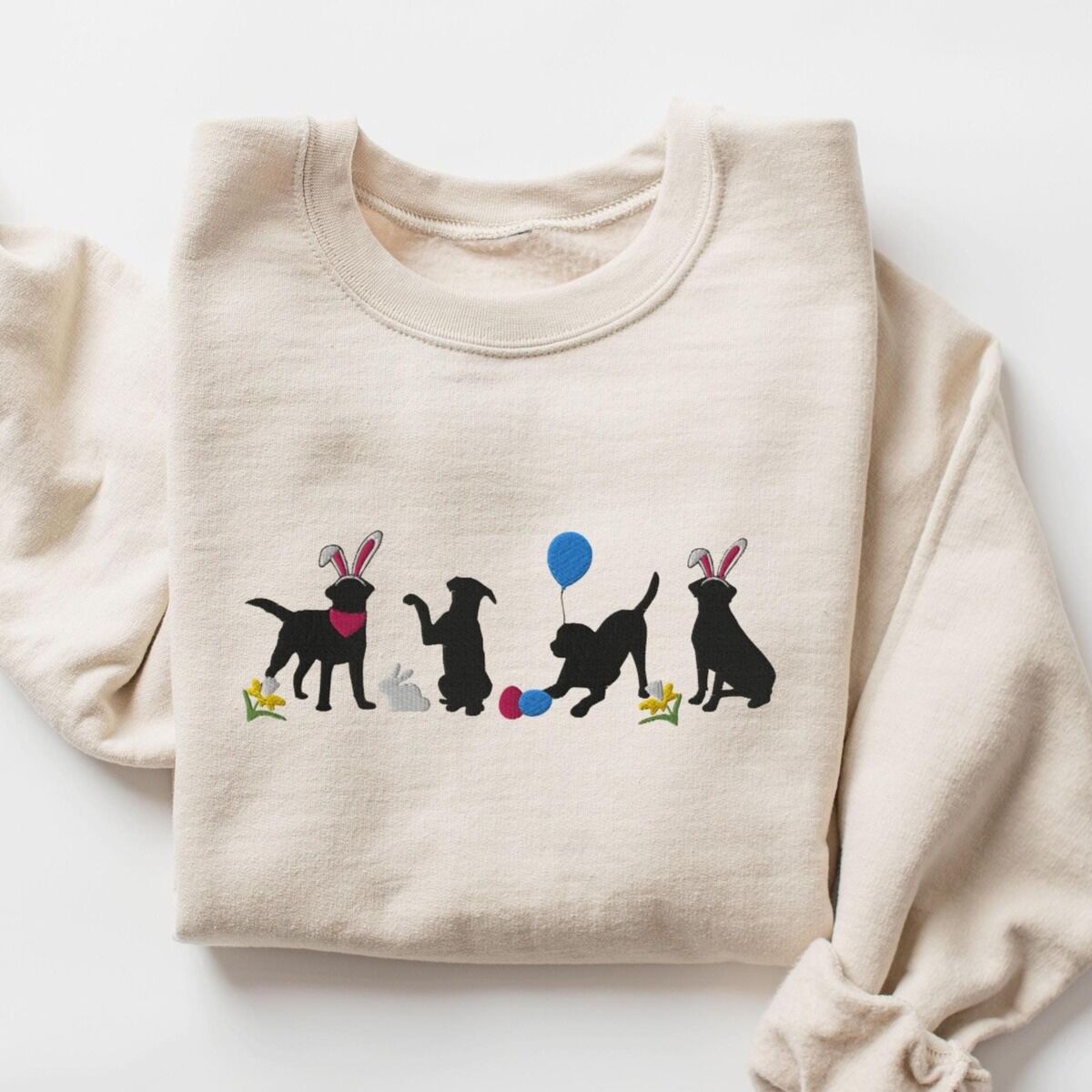 Black Lab Easter Sweatshirt – Sand Sand crewneck sweatshirt laid flat, embroidered with four black lab silhouettes in bunny ears with a little bunny, Easter eggs, daffodils and a blue balloon