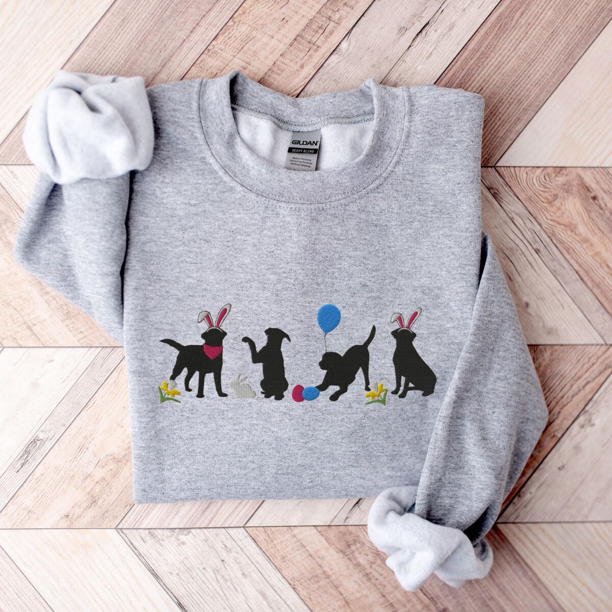 Black Lab Easter Sweatshirt - Sport Grey Sport Grey crewneck sweatshirt laid flat on a wood floor, embroidered with four black lab silhouettes in bunny ears with a little bunny, Easter eggs, daffodils and a blue balloon