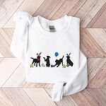 White crewneck sweatshirt laid flat on a wood floor, embroidered with four black lab silhouettes in bunny ears with a little bunny, Easter eggs, daffodils and a blue balloon