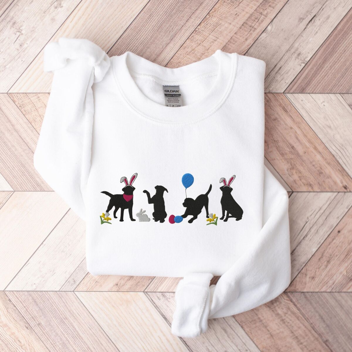 Black Lab Easter Sweatshirt - White White crewneck sweatshirt laid flat on a wood floor, embroidered with four black lab silhouettes in bunny ears with a little bunny, Easter eggs, daffodils and a blue balloon