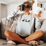 Woman sipping from a mug on a sofa in an Ash crewneck sweatshirt embroidered with three black Labradors, one in an elf hat carrying Christmas lights, one in a candy-cane scarf and one in a Santa hat pawing a gift