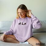 Woman sitting on a sofa in an orchid crewneck sweatshirt embroidered with three black Labradors, one in an elf hat carrying Christmas lights, one in a candy-cane scarf and one in a Santa hat pawing a gift