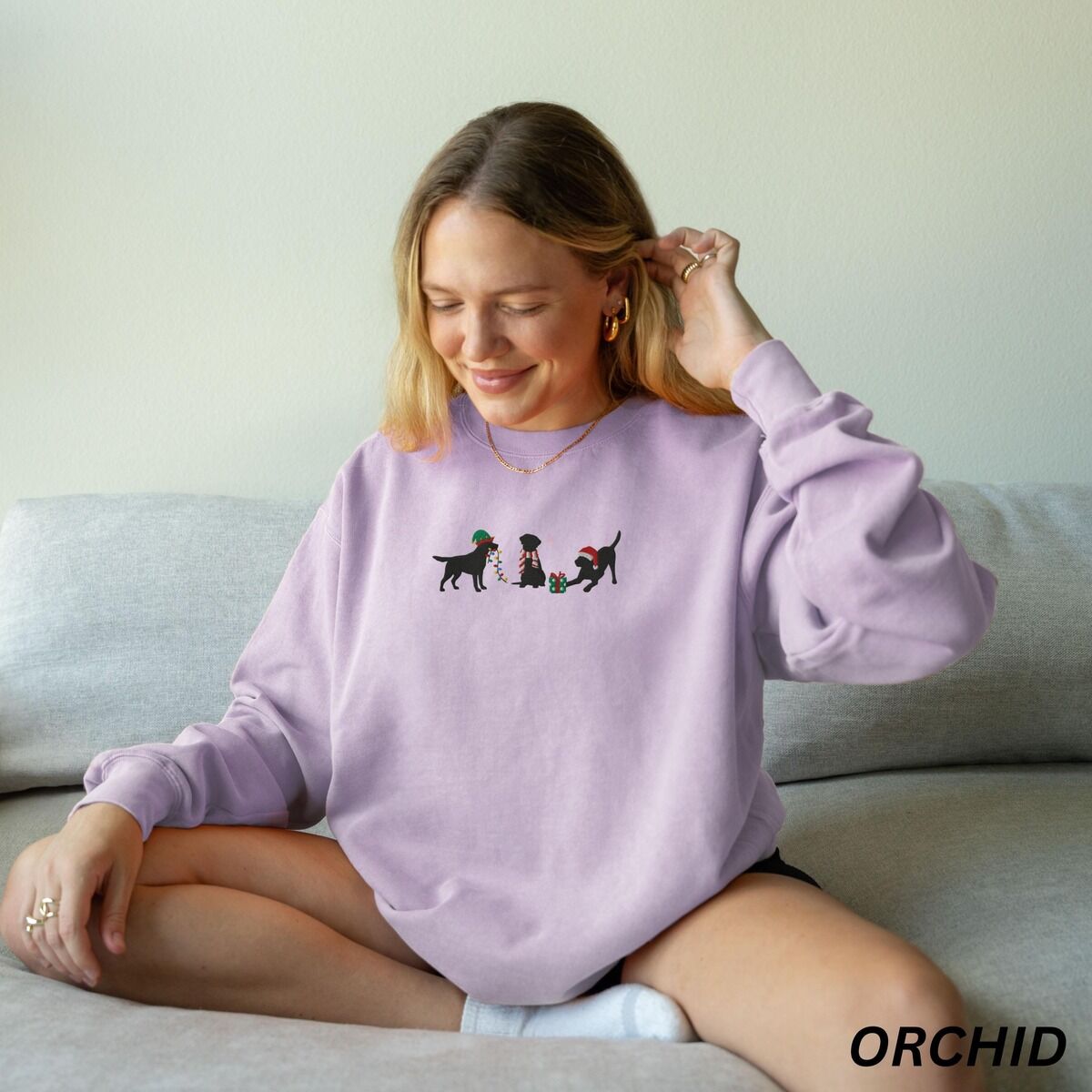 Black Lab Elf Christmas Sweatshirt - worn Woman sitting on a sofa in an orchid crewneck sweatshirt embroidered with three black Labradors, one in an elf hat carrying Christmas lights, one in a candy-cane scarf and one in a Santa hat pawing a gift