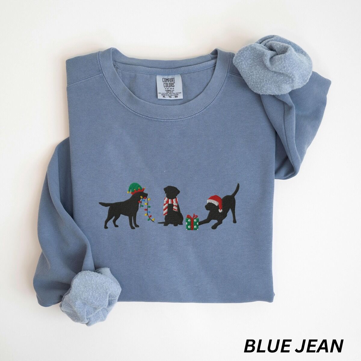 Black Lab Elf Christmas Sweatshirt – Blue Jean Blue Jean crewneck sweatshirt laid flat with rolled cuffs, embroidered with three black Labradors, one in an elf hat carrying Christmas lights, one in a candy-cane scarf and one in a Santa hat pawing a gift