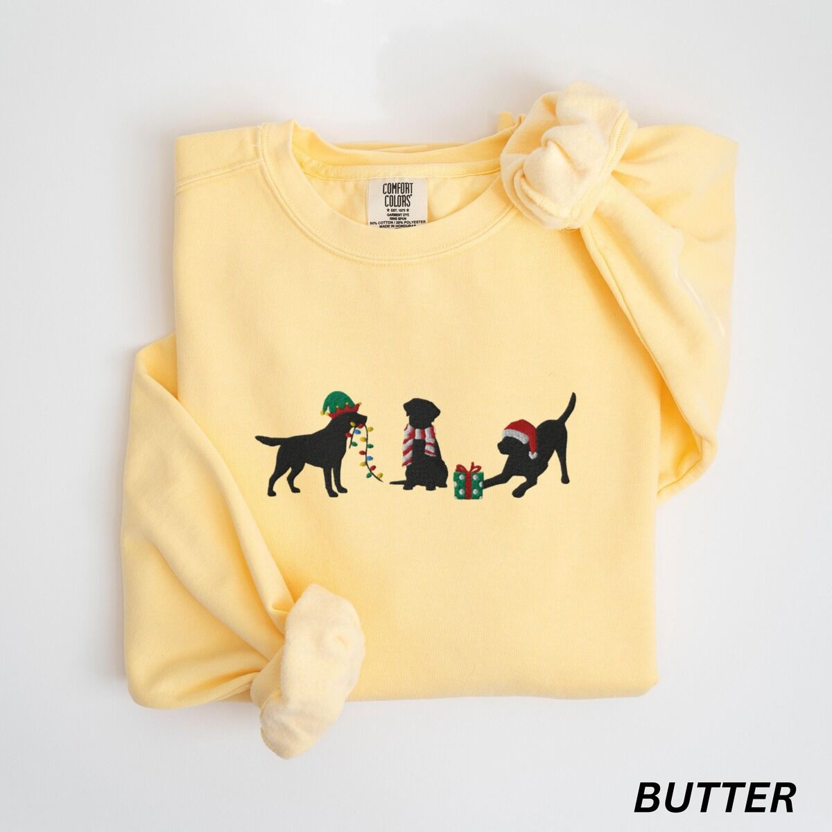 Black Lab Elf Christmas Sweatshirt – Butter Butter crewneck sweatshirt laid flat with rolled cuffs, embroidered with three black Labradors, one in an elf hat carrying Christmas lights, one in a candy-cane scarf and one in a Santa hat pawing a gift