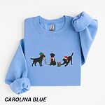 Carolina Blue crewneck sweatshirt laid flat with a rolled cuff, embroidered with three black Labradors, one in an elf hat carrying Christmas lights, one in a candy-cane scarf and one in a Santa hat pawing a gift