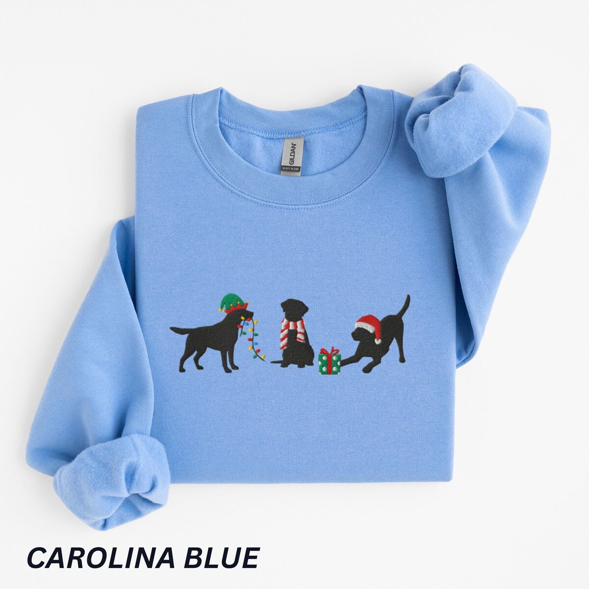 Black Lab Elf Christmas Sweatshirt - Carolina Blue Carolina Blue crewneck sweatshirt laid flat with a rolled cuff, embroidered with three black Labradors, one in an elf hat carrying Christmas lights, one in a candy-cane scarf and one in a Santa hat pawing a gift
