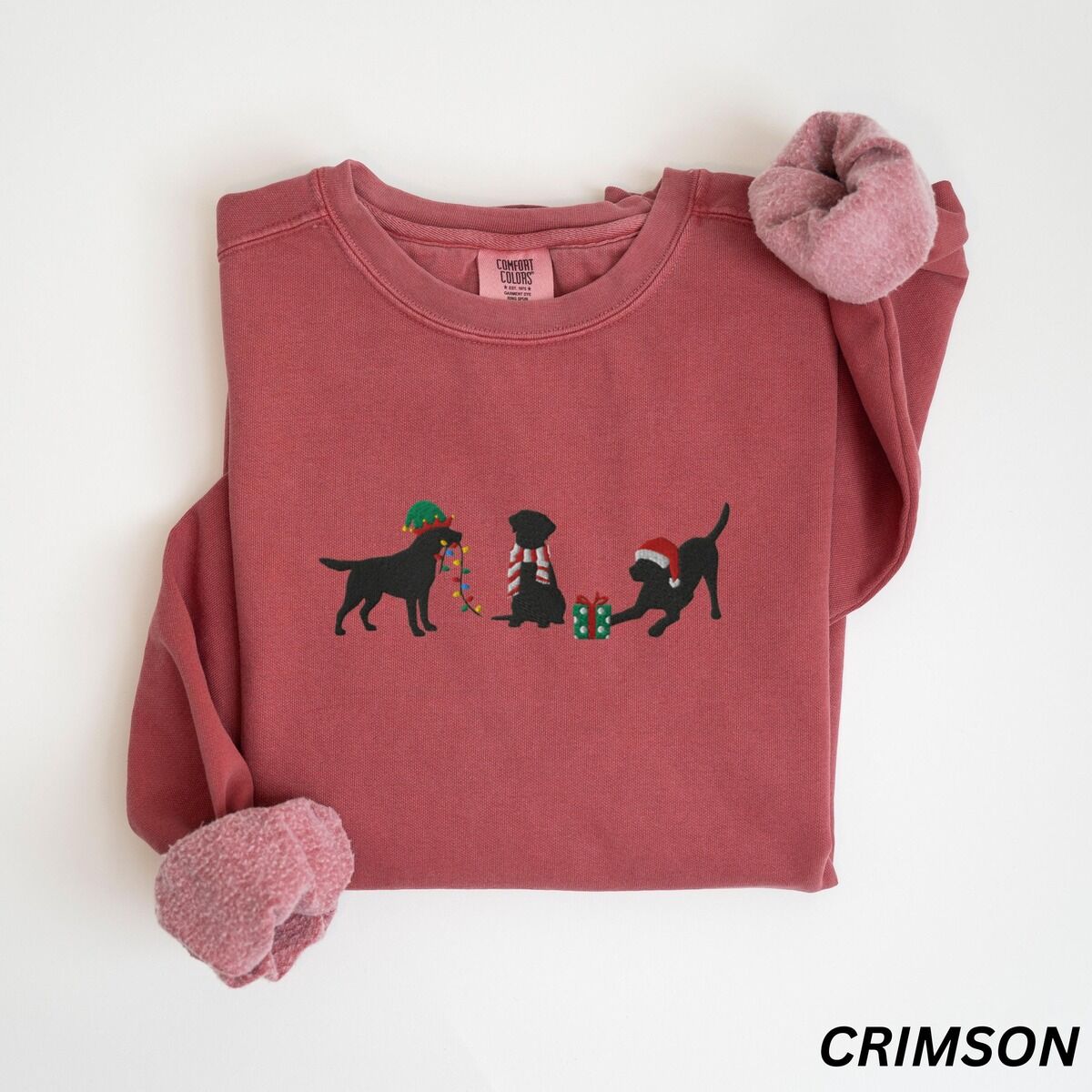 Black Lab Elf Christmas Sweatshirt – Crimson Crimson crewneck sweatshirt laid flat with rolled cuffs, embroidered with three black Labradors, one in an elf hat carrying Christmas lights, one in a candy-cane scarf and one in a Santa hat pawing a gift