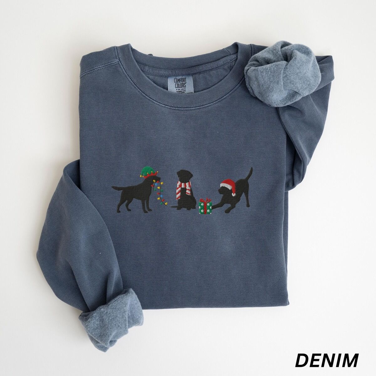 Black Lab Elf Christmas Sweatshirt – Denim Denim crewneck sweatshirt laid flat with rolled cuffs, embroidered with three black Labradors, one in an elf hat carrying Christmas lights, one in a candy-cane scarf and one in a Santa hat pawing a gift