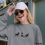 Laughing woman in a cap and sunglasses wearing a Grey crewneck sweatshirt embroidered with three black Labradors, one in an elf hat carrying Christmas lights, one in a candy-cane scarf and one in a Santa hat pawing a gift