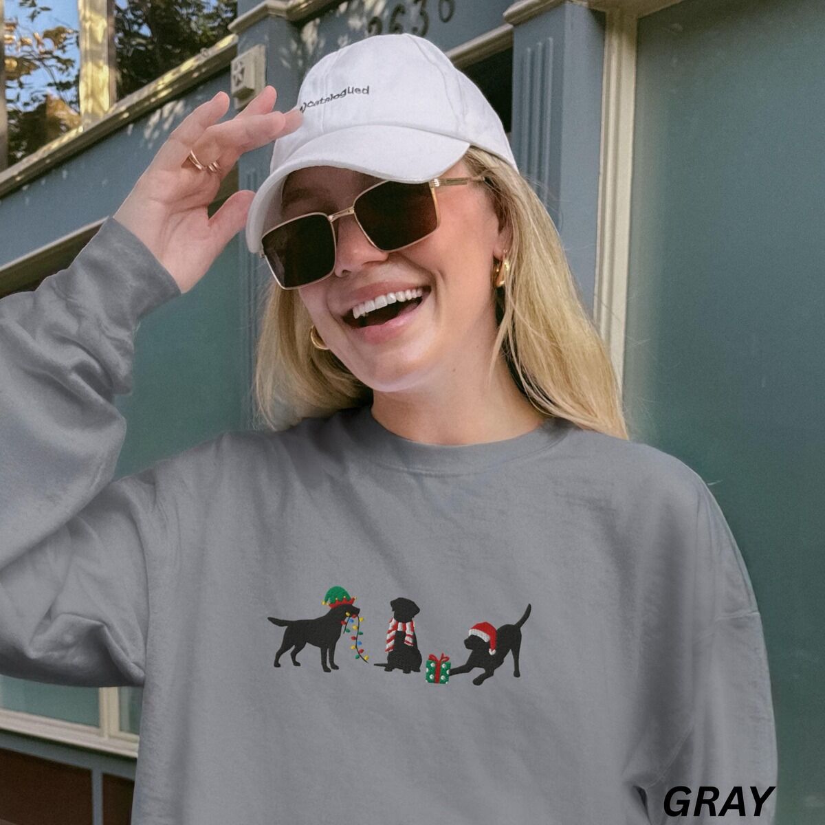 Black Lab Elf Christmas Sweatshirt – Grey Laughing woman in a cap and sunglasses wearing a Grey crewneck sweatshirt embroidered with three black Labradors, one in an elf hat carrying Christmas lights, one in a candy-cane scarf and one in a Santa hat pawing a gift