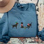 Indigo Blue crewneck sweatshirt laid flat beside a hat and pampas grass, embroidered with three black Labradors, one in an elf hat carrying Christmas lights, one in a candy-cane scarf and one in a Santa hat pawing a gift