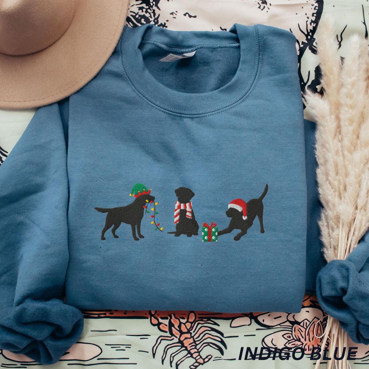 Black Lab Elf Christmas Sweatshirt - Indigo Blue Indigo Blue crewneck sweatshirt laid flat beside a hat and pampas grass, embroidered with three black Labradors, one in an elf hat carrying Christmas lights, one in a candy-cane scarf and one in a Santa hat pawing a gift