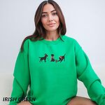 Woman sitting in an Irish Green crewneck sweatshirt embroidered with three black Labradors, one in an elf hat carrying Christmas lights, one in a candy-cane scarf and one in a Santa hat pawing a gift