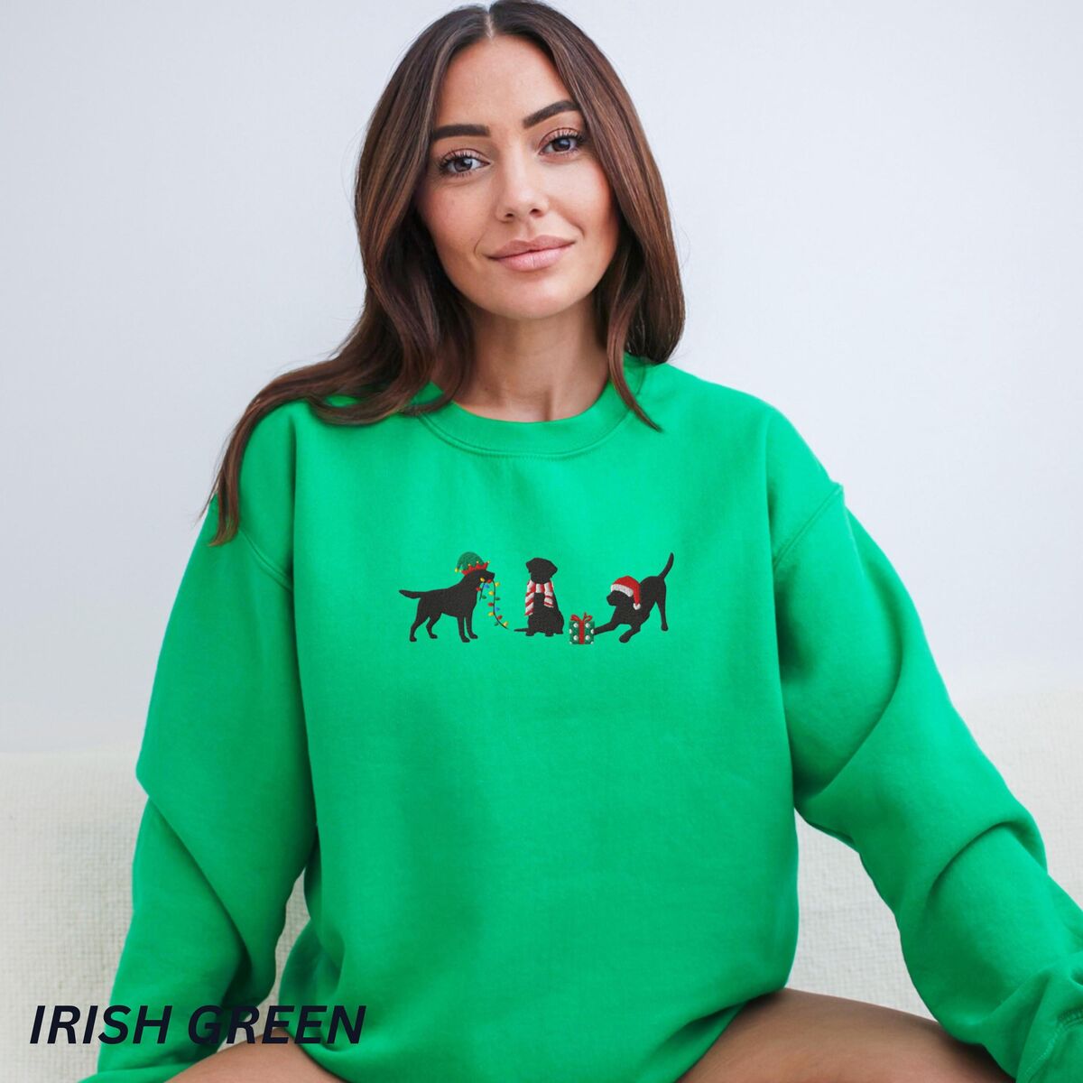 Black Lab Elf Christmas Sweatshirt - Irish Green Woman sitting in an Irish Green crewneck sweatshirt embroidered with three black Labradors, one in an elf hat carrying Christmas lights, one in a candy-cane scarf and one in a Santa hat pawing a gift