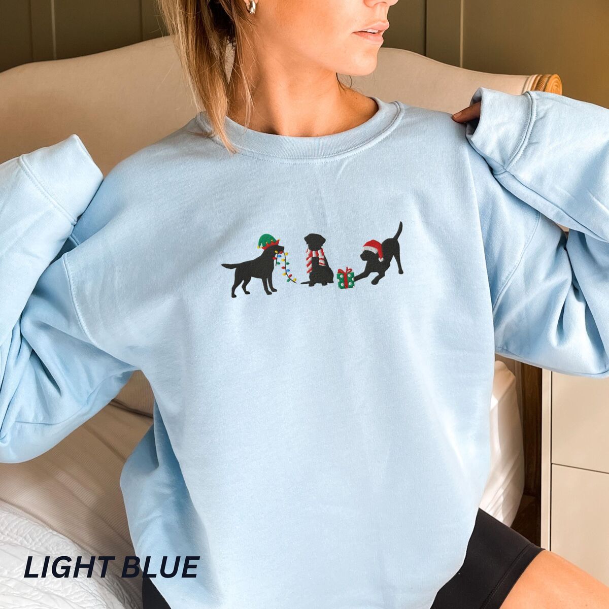 Black Lab Elf Christmas Sweatshirt - Light Blue Woman in a Light Blue crewneck sweatshirt embroidered with three black Labradors, one in an elf hat carrying Christmas lights, one in a candy-cane scarf and one in a Santa hat pawing a gift