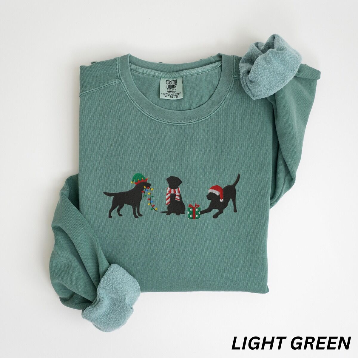 Black Lab Elf Christmas Sweatshirt – Light Green Light Green crewneck sweatshirt laid flat with rolled cuffs, embroidered with three black Labradors, one in an elf hat carrying Christmas lights, one in a candy-cane scarf and one in a Santa hat pawing a gift
