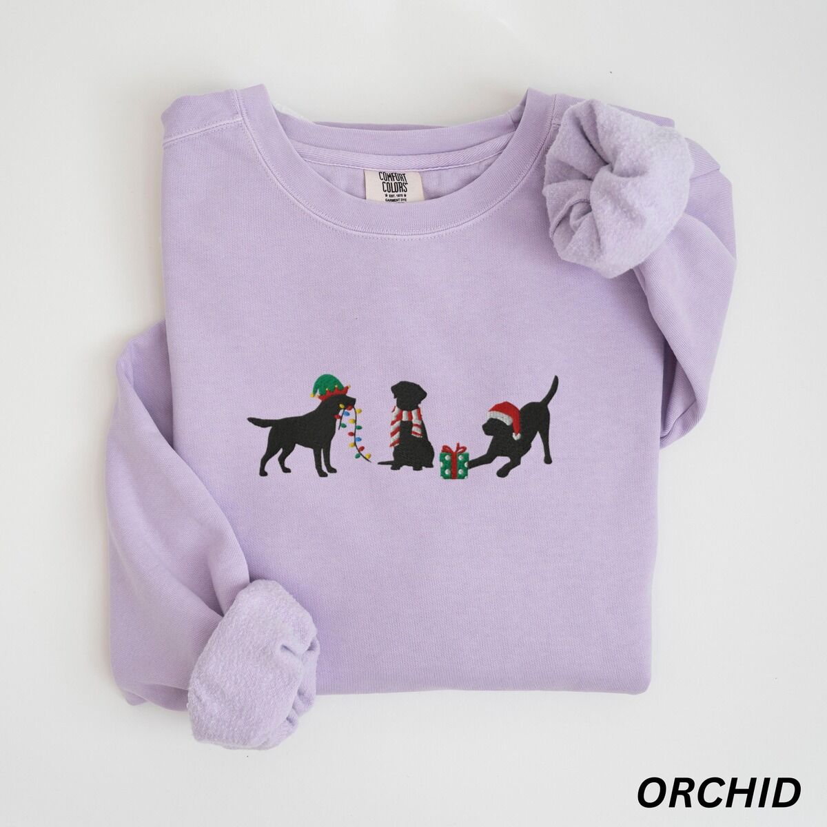 Black Lab Elf Christmas Sweatshirt – Orchid Orchid crewneck sweatshirt laid flat with rolled cuffs, embroidered with three black Labradors, one in an elf hat carrying Christmas lights, one in a candy-cane scarf and one in a Santa hat pawing a gift