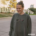 Woman outdoors on a street in a Pepper crewneck sweatshirt embroidered with three black Labradors, one in an elf hat carrying Christmas lights, one in a candy-cane scarf and one in a Santa hat pawing a gift