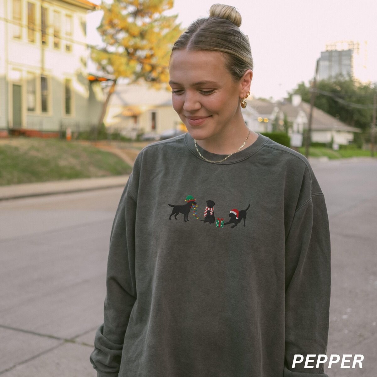 Black Lab Elf Christmas Sweatshirt – Pepper Woman outdoors on a street in a Pepper crewneck sweatshirt embroidered with three black Labradors, one in an elf hat carrying Christmas lights, one in a candy-cane scarf and one in a Santa hat pawing a gift