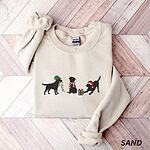 Sand crewneck sweatshirt laid flat on a chevron wood floor, embroidered with three black Labradors, one in an elf hat carrying Christmas lights, one in a candy-cane scarf and one in a Santa hat pawing a gift