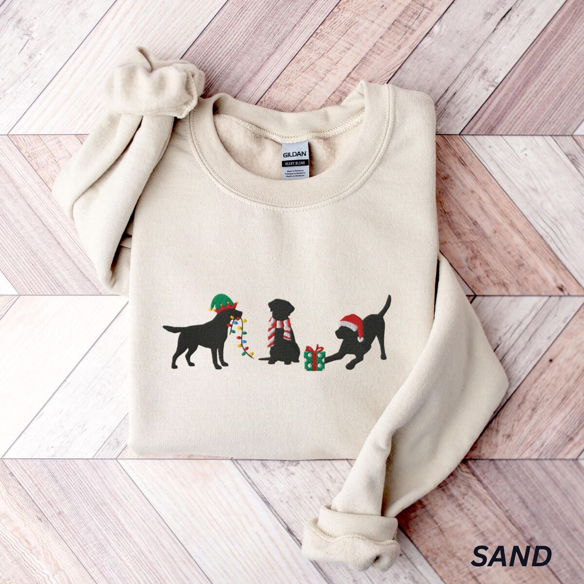 Black Lab Elf Christmas Sweatshirt - Sand Sand crewneck sweatshirt laid flat on a chevron wood floor, embroidered with three black Labradors, one in an elf hat carrying Christmas lights, one in a candy-cane scarf and one in a Santa hat pawing a gift