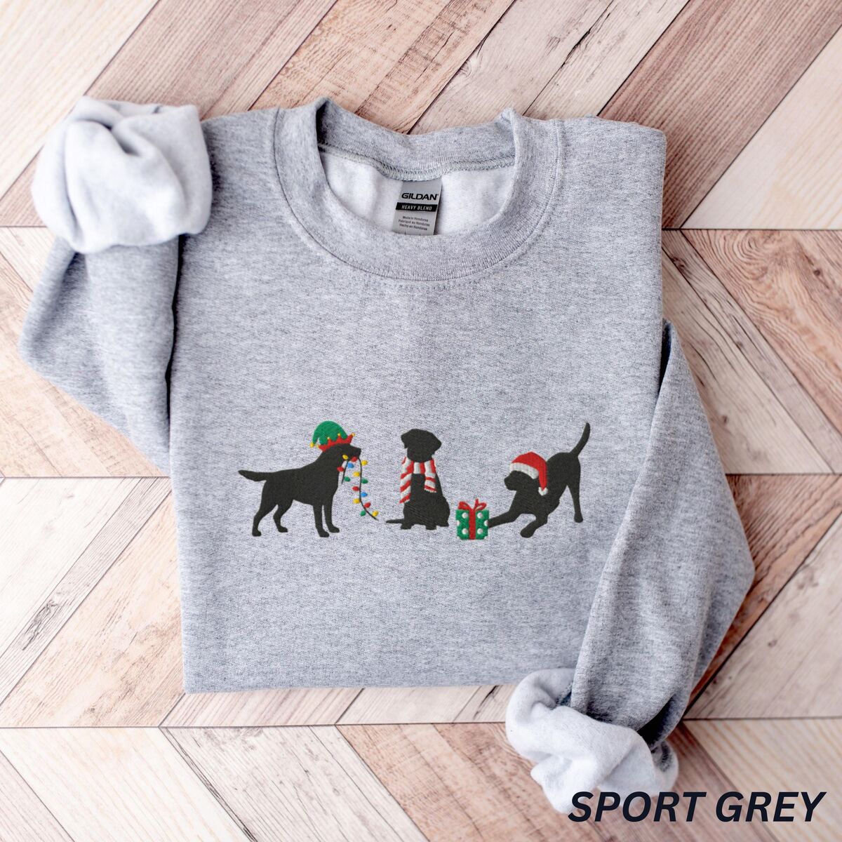 Black Lab Elf Christmas Sweatshirt - Sport Grey Sport Grey crewneck sweatshirt laid flat on a chevron wood floor, embroidered with three black Labradors, one in an elf hat carrying Christmas lights, one in a candy-cane scarf and one in a Santa hat pawing a gift