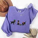 Violet crewneck sweatshirt laid flat beside a hat and a dried palm leaf, embroidered with three black Labradors, one in an elf hat carrying Christmas lights, one in a candy-cane scarf and one in a Santa hat pawing a gift