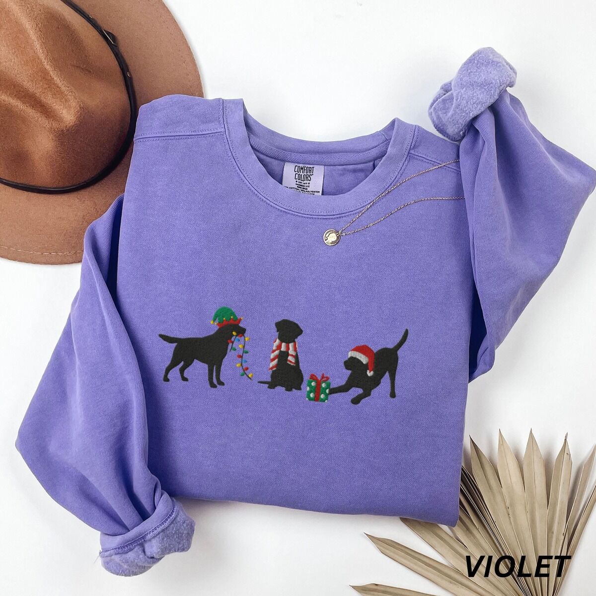 Black Lab Elf Christmas Sweatshirt – Violet Violet crewneck sweatshirt laid flat beside a hat and a dried palm leaf, embroidered with three black Labradors, one in an elf hat carrying Christmas lights, one in a candy-cane scarf and one in a Santa hat pawing a gift