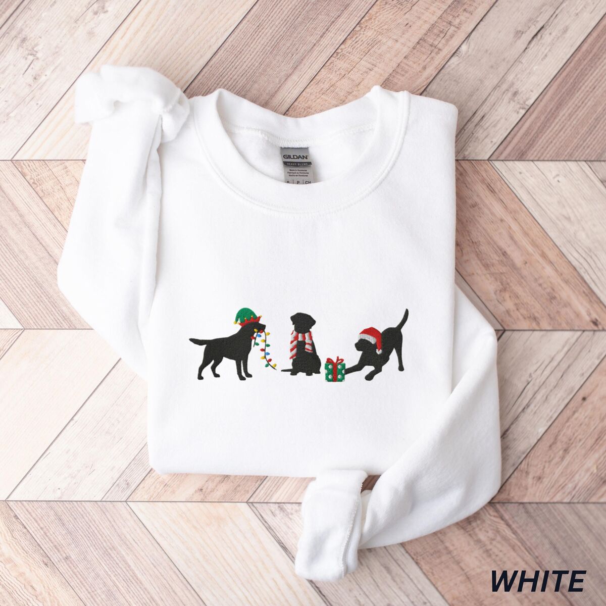 Black Lab Elf Christmas Sweatshirt - White White crewneck sweatshirt laid flat on a chevron wood floor, embroidered with three black Labradors, one in an elf hat carrying Christmas lights, one in a candy-cane scarf and one in a Santa hat pawing a gift