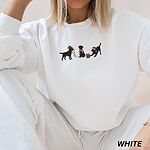 Woman sitting in a White crewneck sweatshirt embroidered with three black Labradors, one in an elf hat carrying Christmas lights, one in a candy-cane scarf and one in a Santa hat pawing a gift