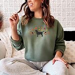 Woman on a sofa in a Military Green crewneck sweatshirt embroidered with a black Labrador silhouette under an arc of orange, red and gold autumn leaves across the chest