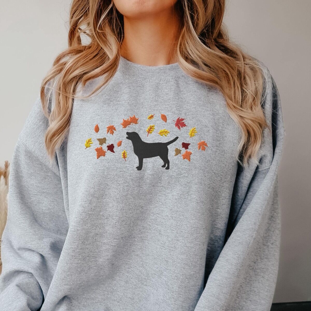 Black Lab Fall Sweatshirt - worn in grey Woman in a Sport Grey crewneck sweatshirt embroidered with a black Labrador silhouette under an arc of orange, red and gold autumn leaves across the chest