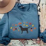 Indigo Blue crewneck sweatshirt laid flat with a hat and pampas grass, embroidered with a black Labrador silhouette under an arc of orange, red and gold autumn leaves across the chest