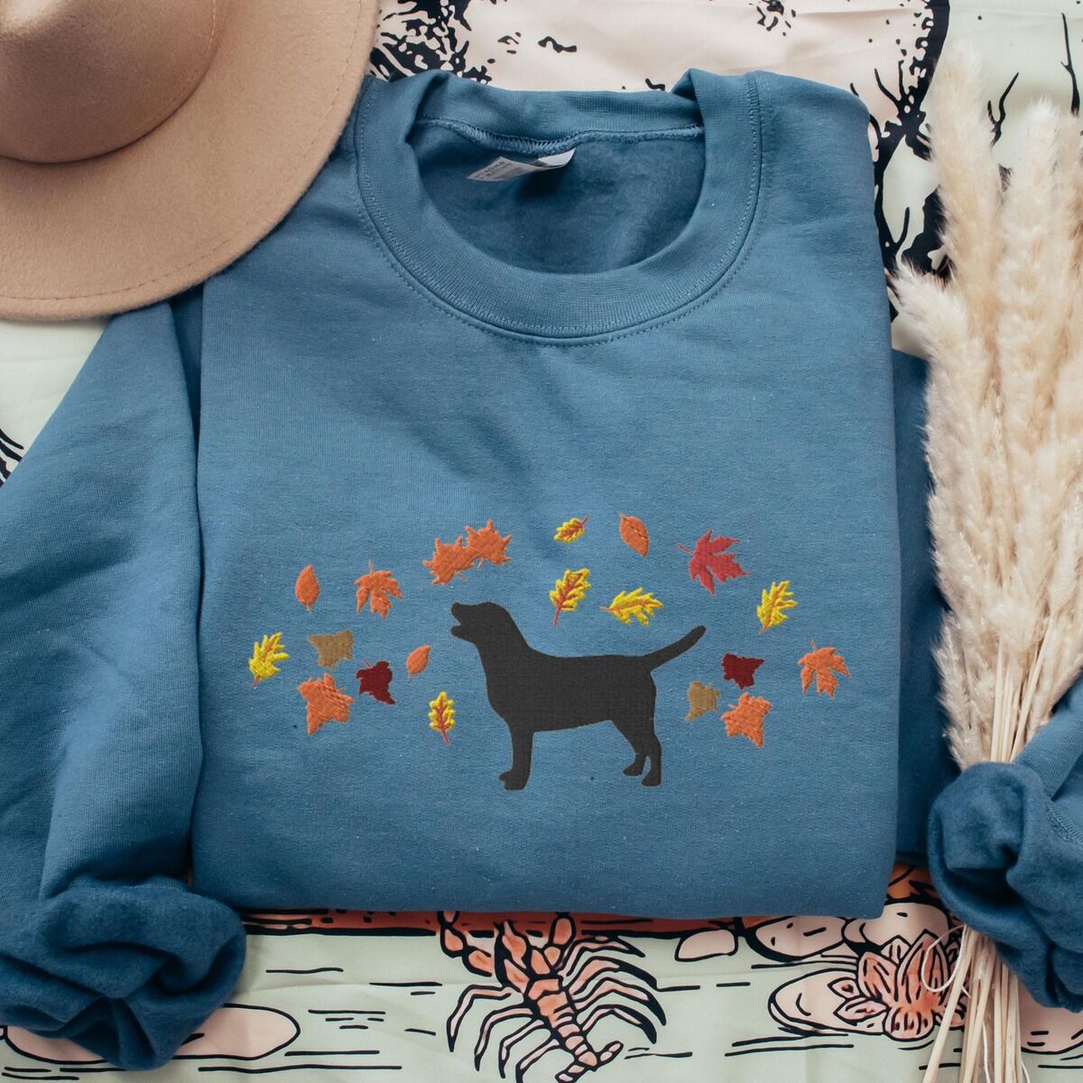 Black Lab Fall Sweatshirt – Indigo Blue Indigo Blue crewneck sweatshirt laid flat with a hat and pampas grass, embroidered with a black Labrador silhouette under an arc of orange, red and gold autumn leaves across the chest