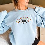 Woman in a Light Blue crewneck sweatshirt embroidered with a black Labrador silhouette under an arc of orange, red and gold autumn leaves across the chest