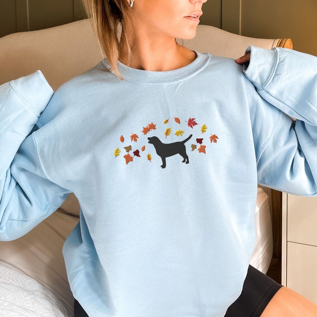 Black Lab Fall Sweatshirt - Light Blue Woman in a Light Blue crewneck sweatshirt embroidered with a black Labrador silhouette under an arc of orange, red and gold autumn leaves across the chest